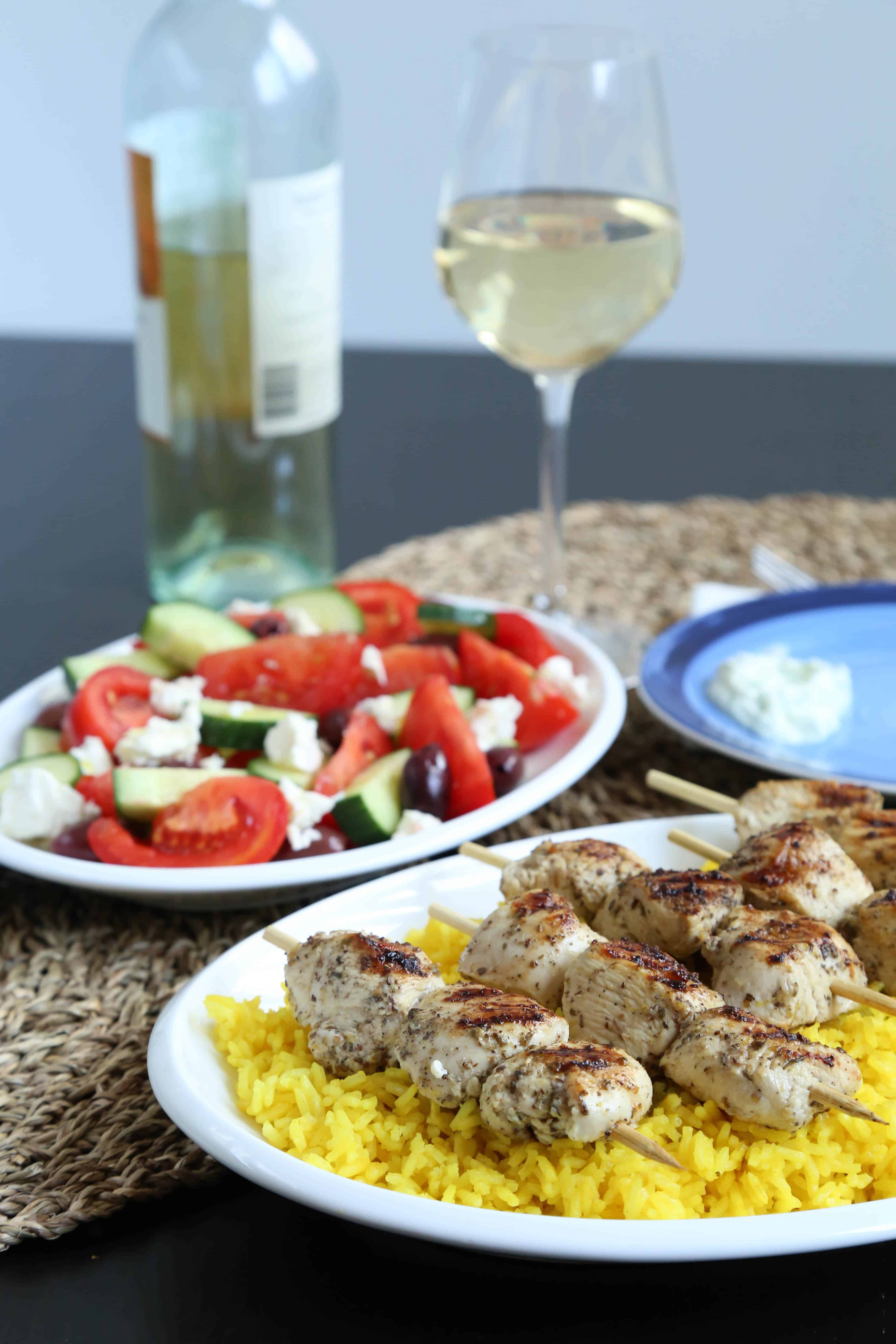 Chicken kabobs, rice, Greek salad, wine glasses, and a bottle of wine on a dinner table.