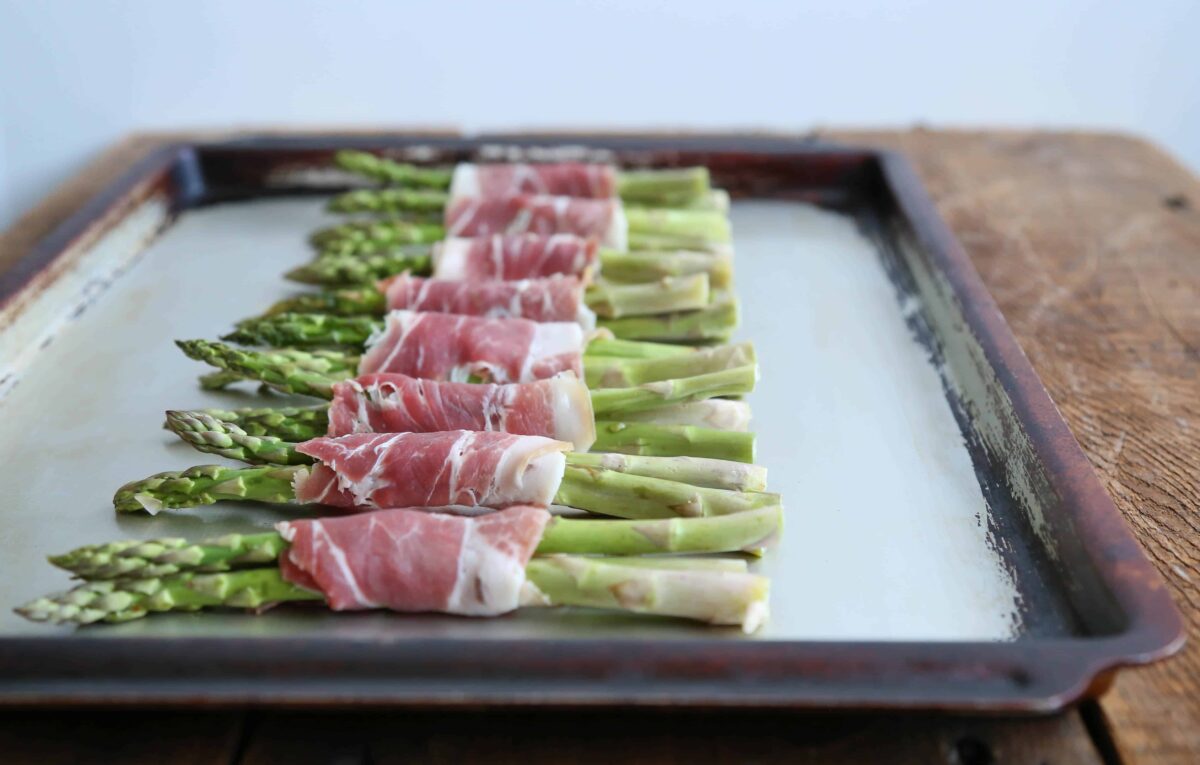 uncooked prosciutto wrapped asparagus lined up on a sheet pan.