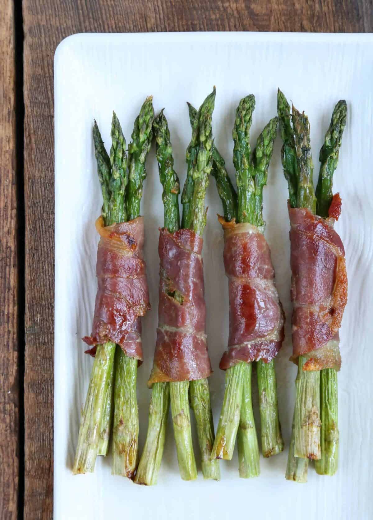 prosciutto wrapped asparagus on a white platter sitting on a wooden table.