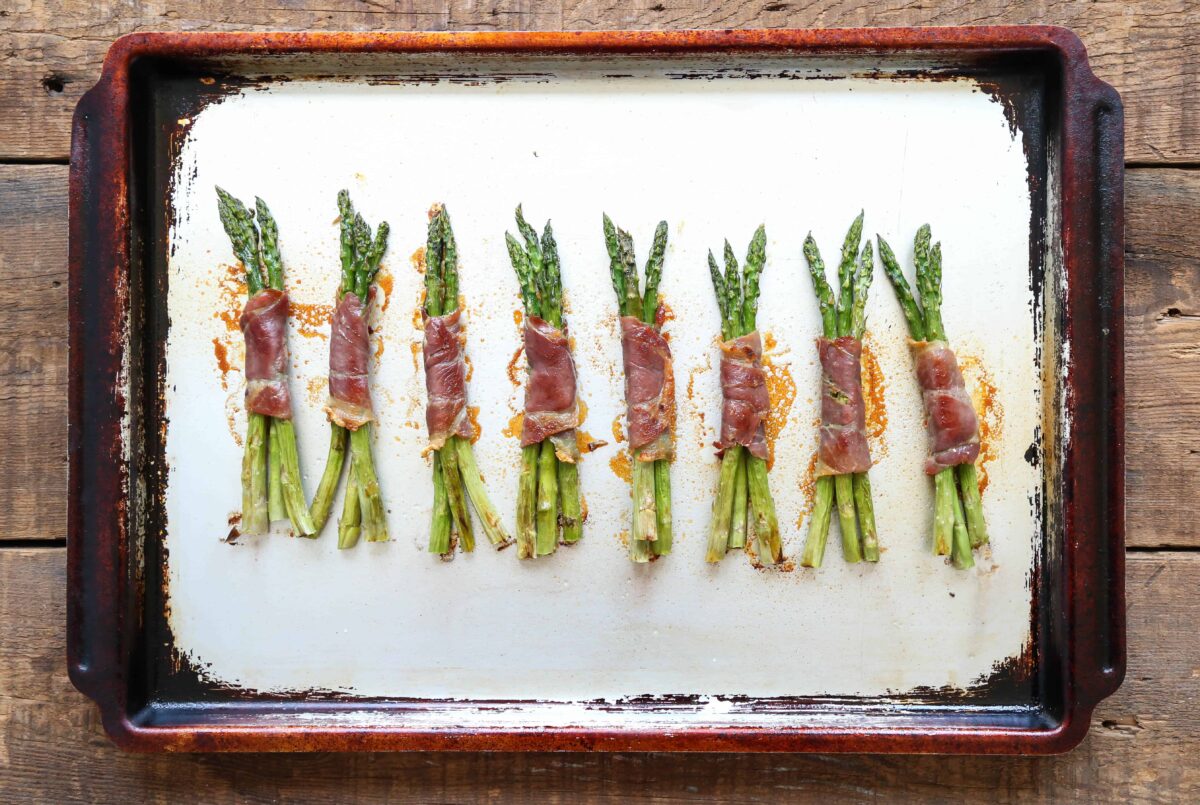 prosciutto wrapped asparagus lined up on a metal sheet tray.