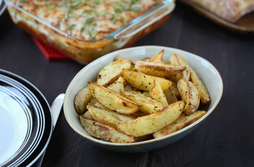 crispy garlic potatoes in a beige bowl with a glass pan of lasagna in the background.