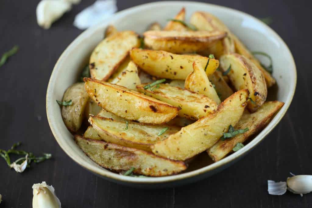 crispy garlic potatoes in a beige bowl surrounded by garlic cloves on a brown table.