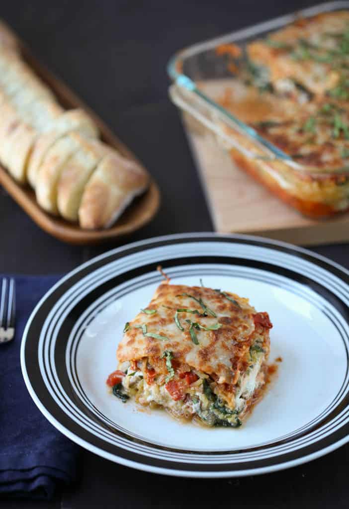 piece of spaghetti squash lasagna on a black and white plate with a whole lasagna and sliced bread in the background.