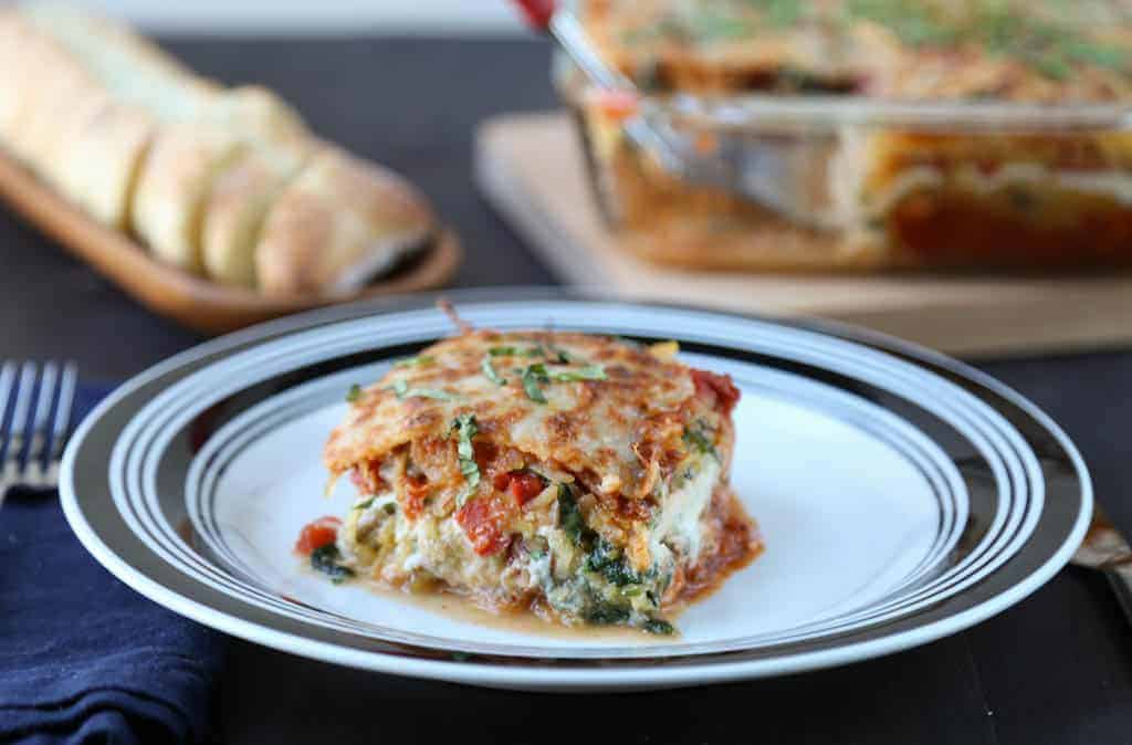 piece of spaghetti squash lasagna on a black and white plate with a whole lasagna and sliced bread in the background.