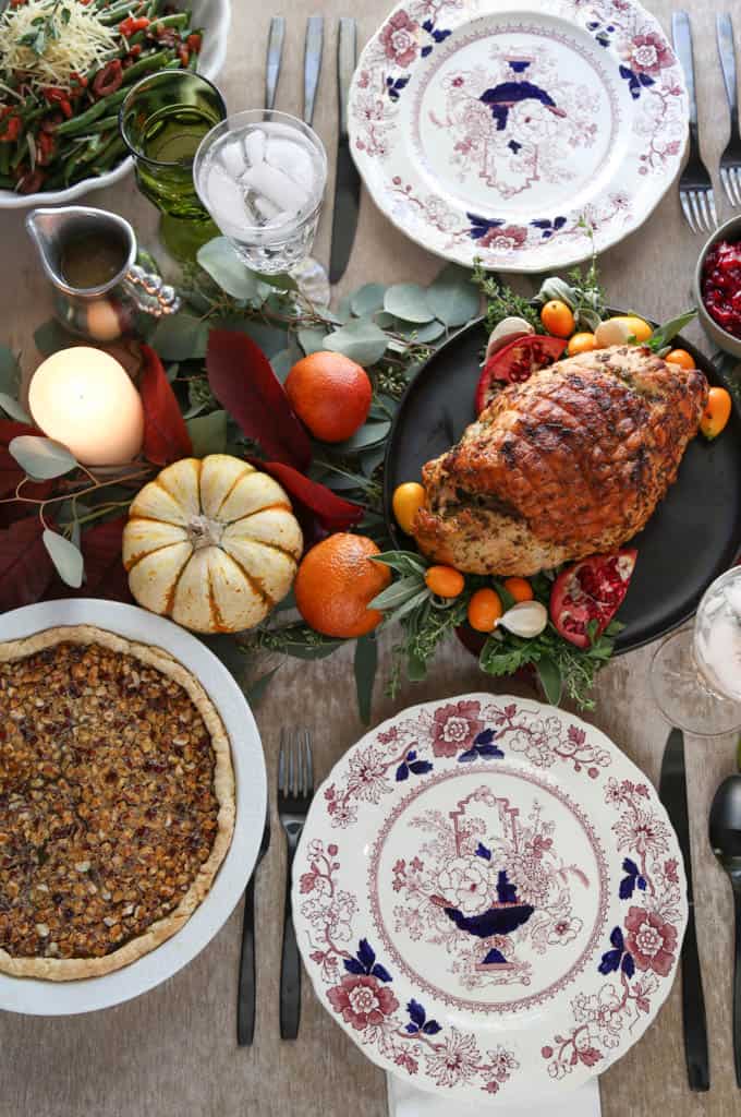 table set for thanksgiving with a turkey breast, pumpkins, a hazelnut pie, and decorative purple and white china plates.