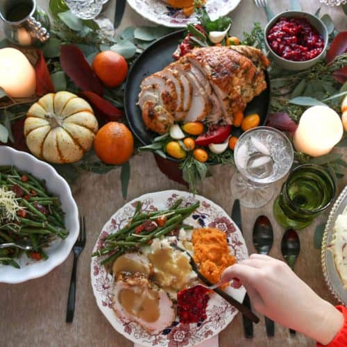 hand scooping cranberry sauce onto a loaded thanksgiving plate sitting on a table decorated for thanksgiving.