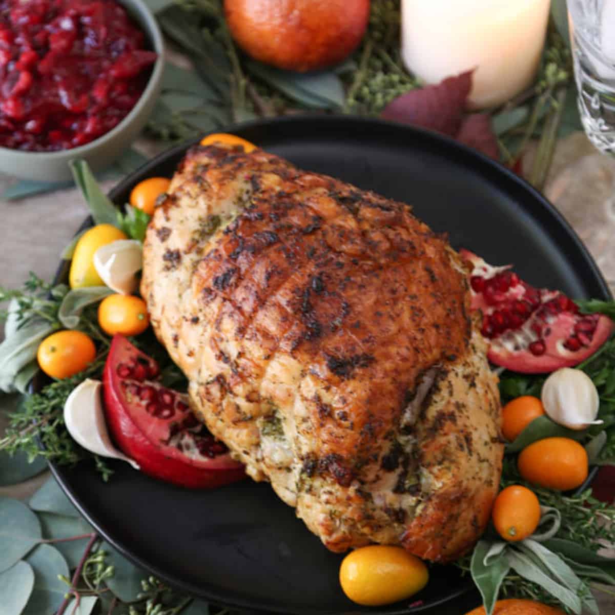 cooked turkey breast on a black platter with pomegranates, herbs, and kumquats for garnish.