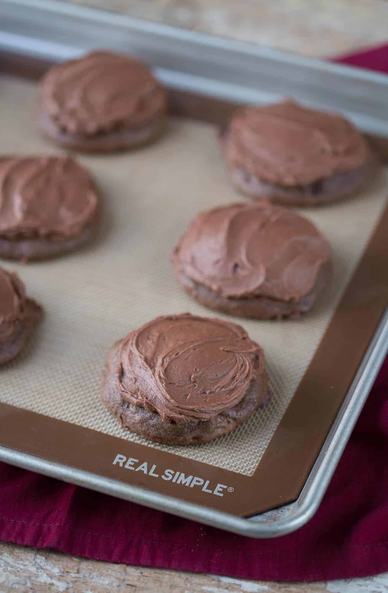 chocolate cookies with chocolate frosting on a sheet pan, sitting on a red towel.