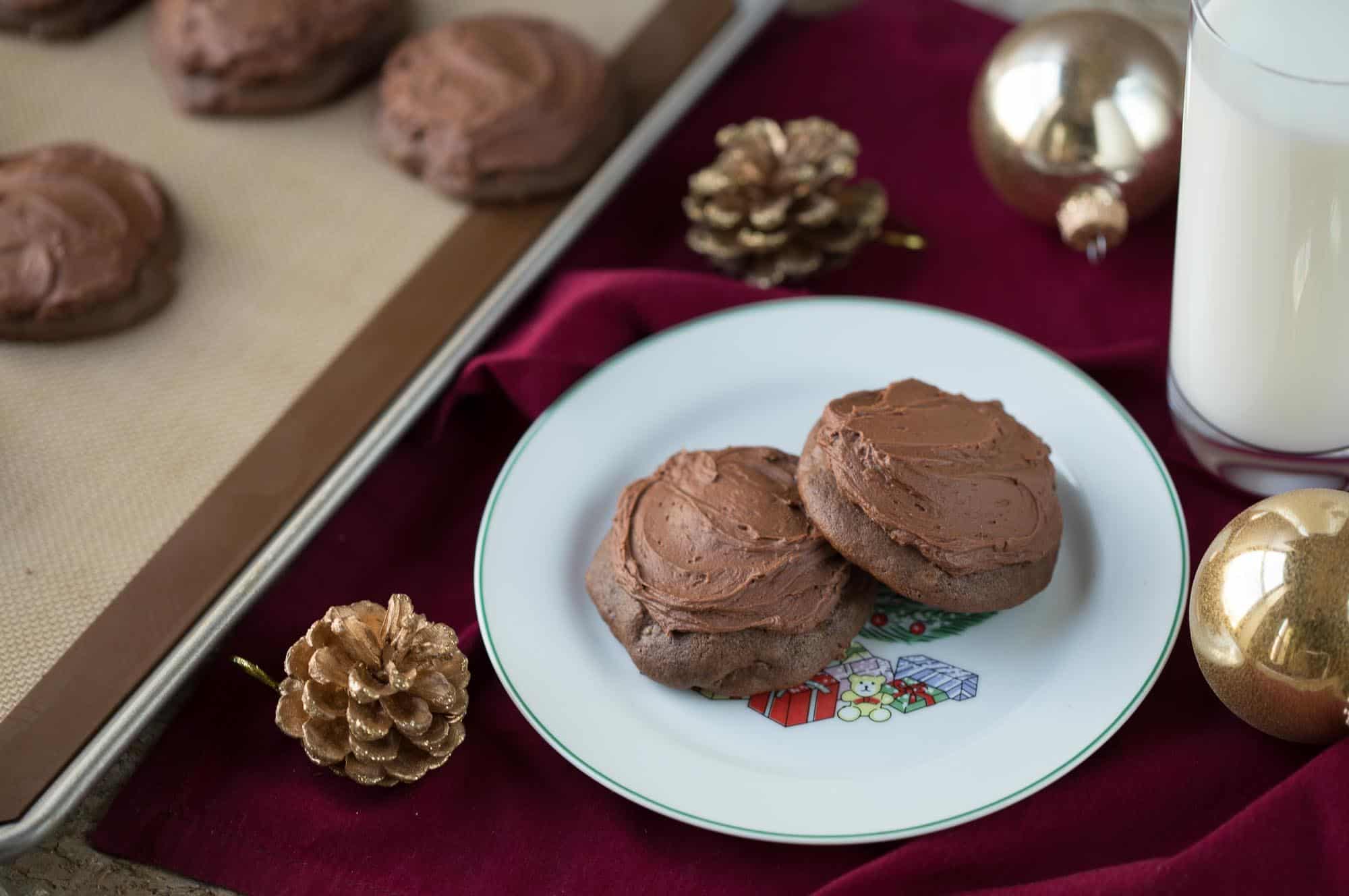 chocolate cookies with chocolate frosting on a white plate surrounded by gold pinecones on a deep red tablecloth.