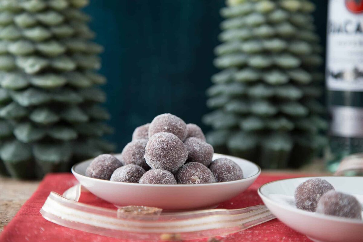 a tower of rum balls in a white dish with green wax christmas trees behind it.