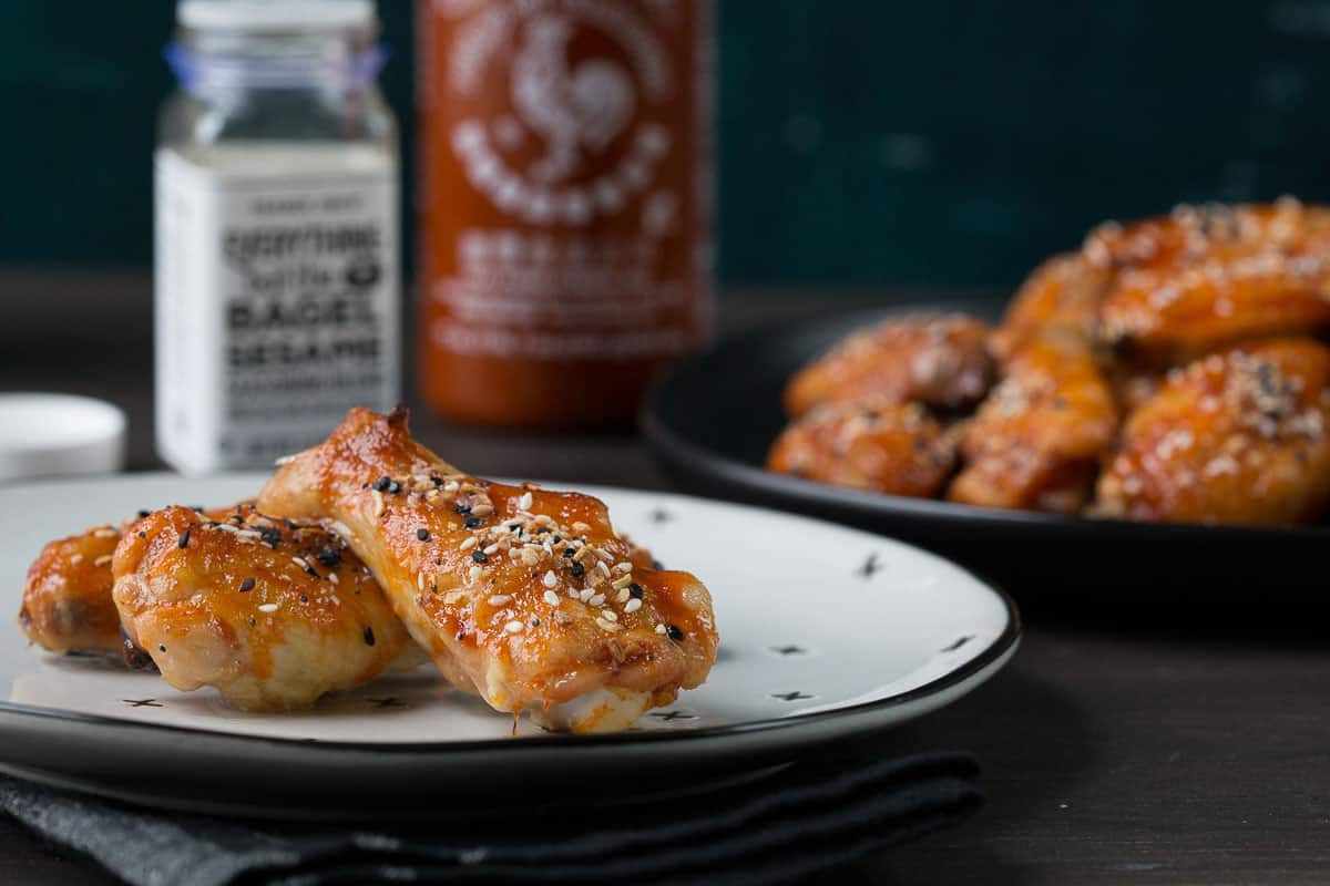 baked chicken wings on a white plate next to a bottle of sriracha and a jar of everything bagel seasoning.