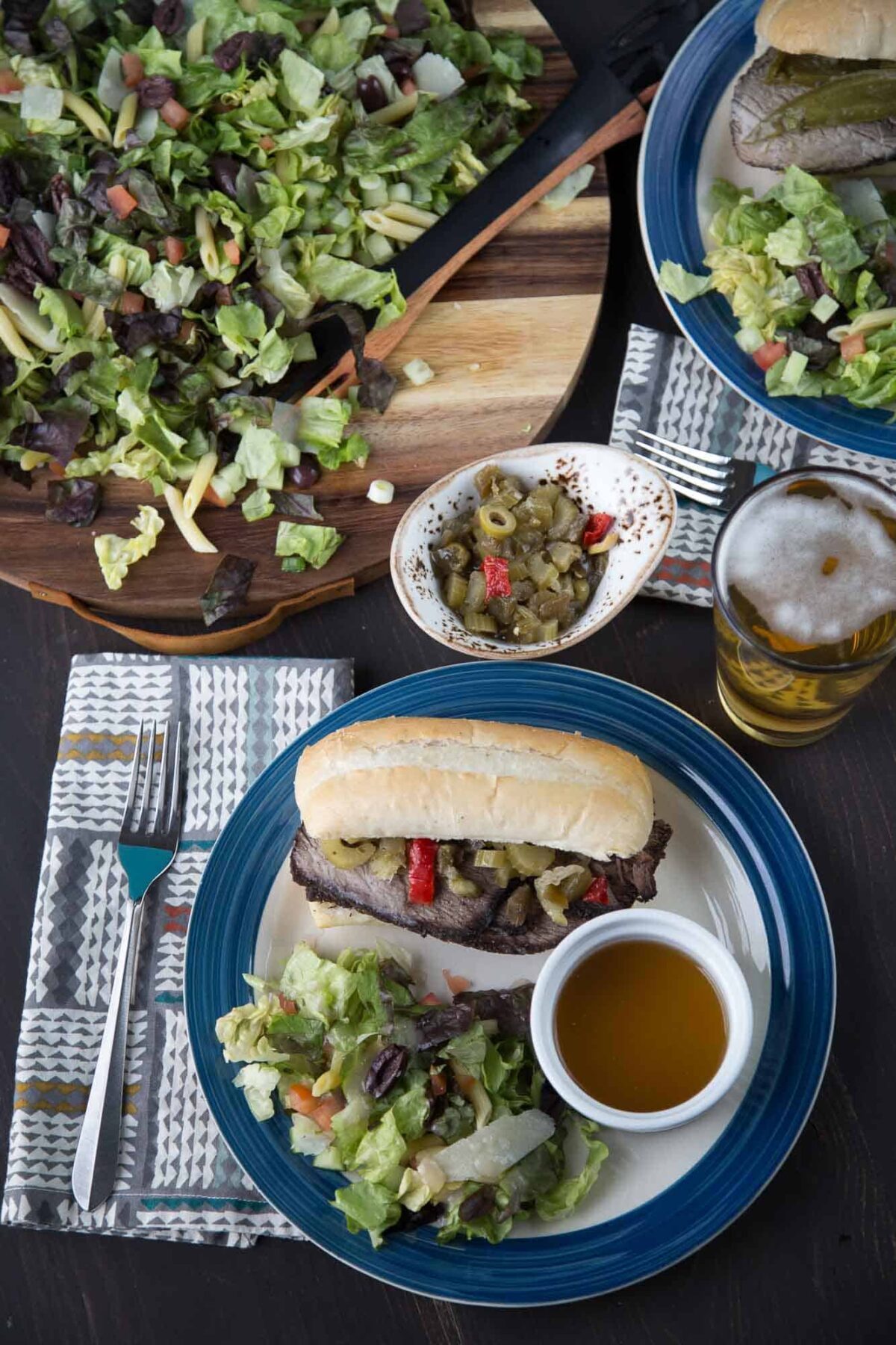 dinner table with italian beef sandwiches, a leafy green salad, and sides of giardiniera and au jus.