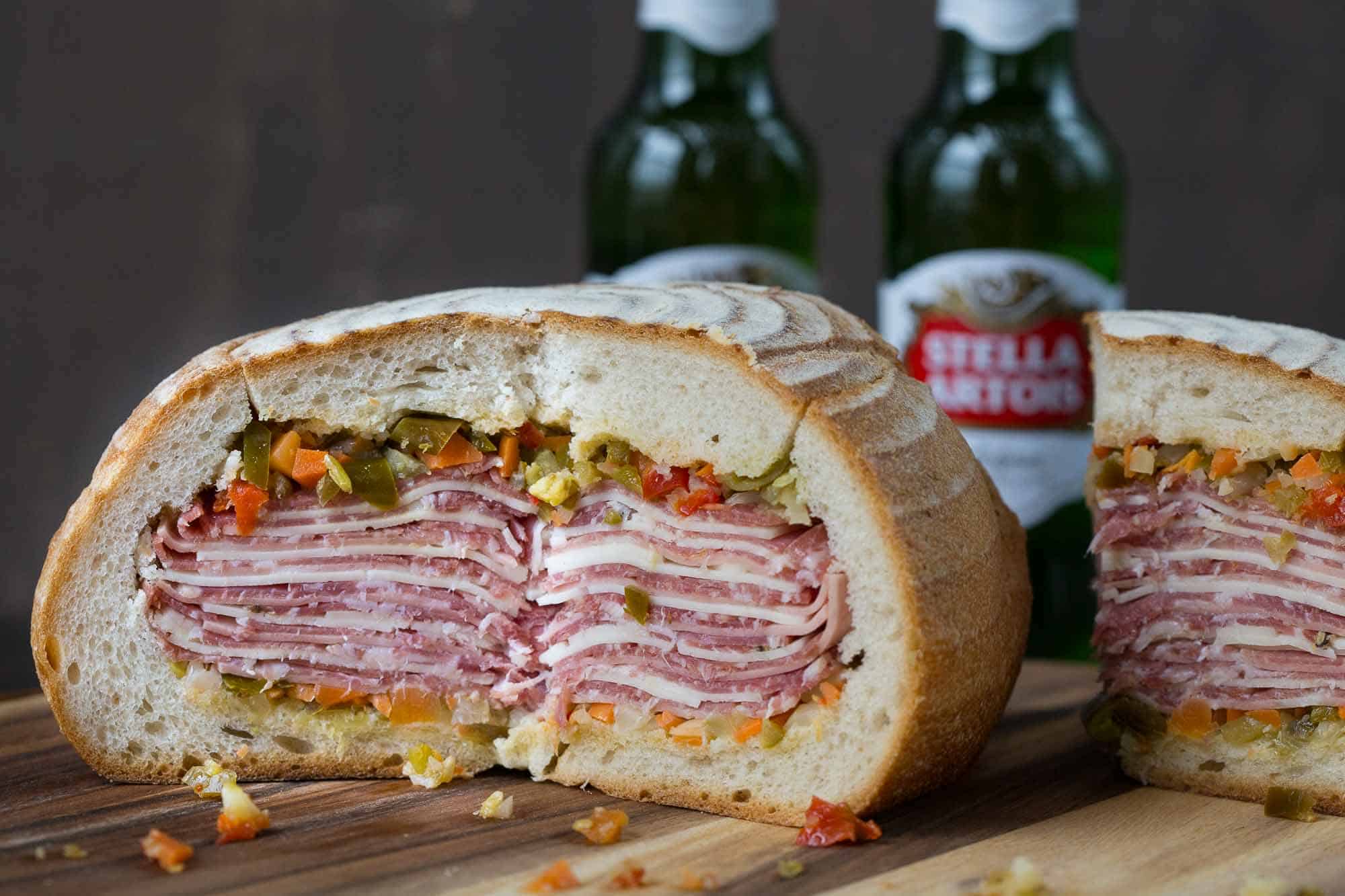 gigantic muffaletta cut open with bottles of beer in the background.