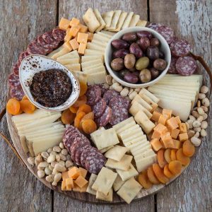 round cheese board with sliced cheese, salami, nuts, dried fruit, and little bowls of jam and olives.