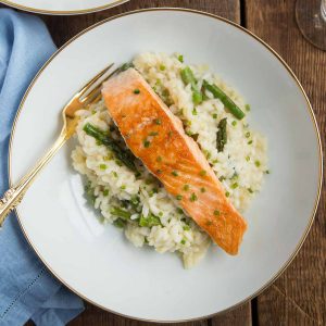 piece of seared salmon on top of asparagus risotto in a white shallow bowl with a gold fork.