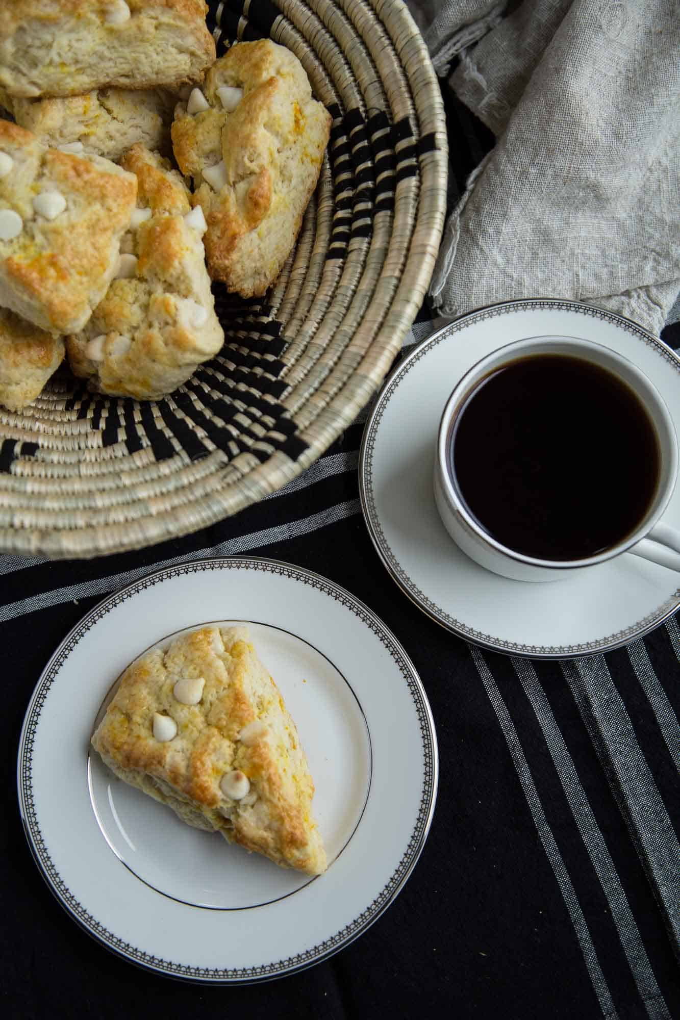 single scone on a plate next to a basket of scones and a teacup of coffee.