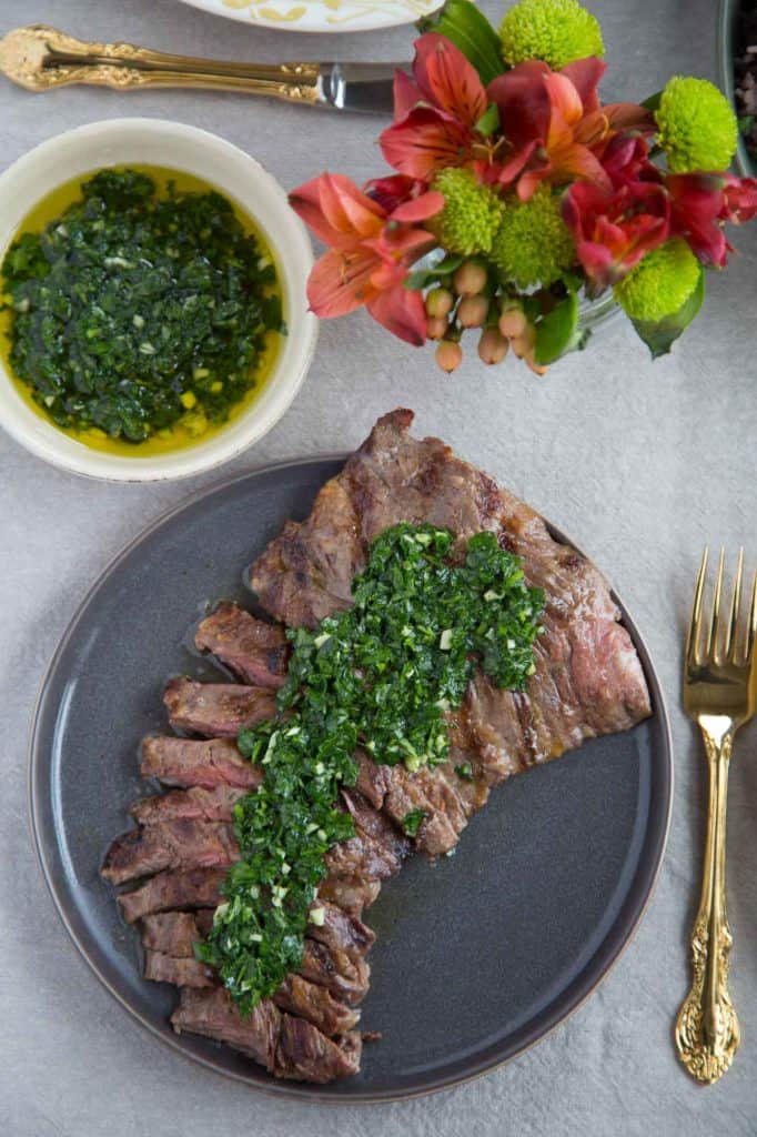 skirt steak sliced on a gray platter with chimichurri sauce on top, next to a bowl of more chimichurri and a small bouquet of fresh flowers.