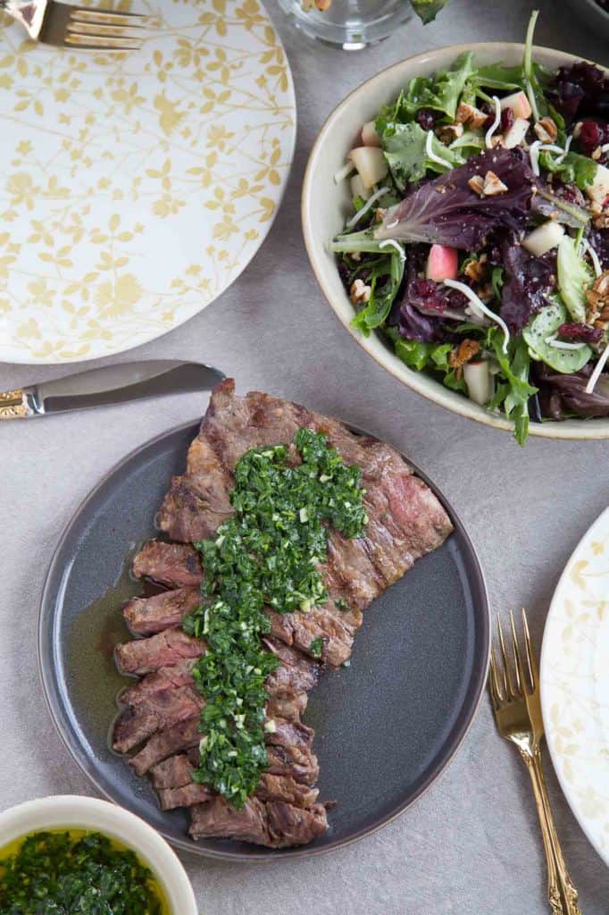 grilled skirt steak on a gray plate topped with chimichurri, next to a big lettuce salad.
