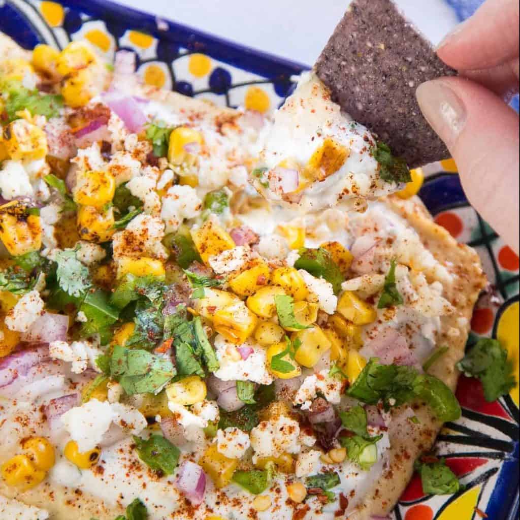 Tortilla chip scooping up mexican street corn dip from a colorful platter.