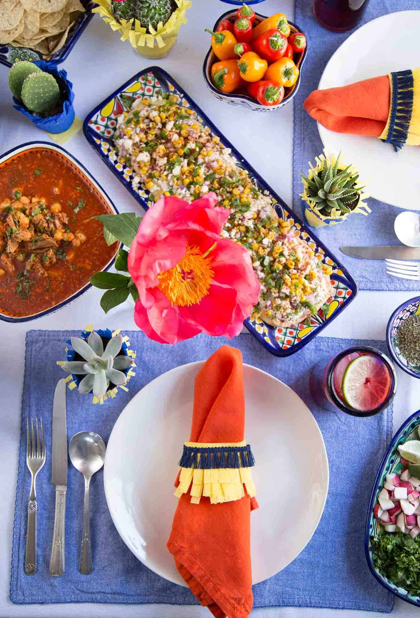 table set with white plates, silverware, an orange napkin with a colorful napkin ring, mexican food, and a single pink peony.