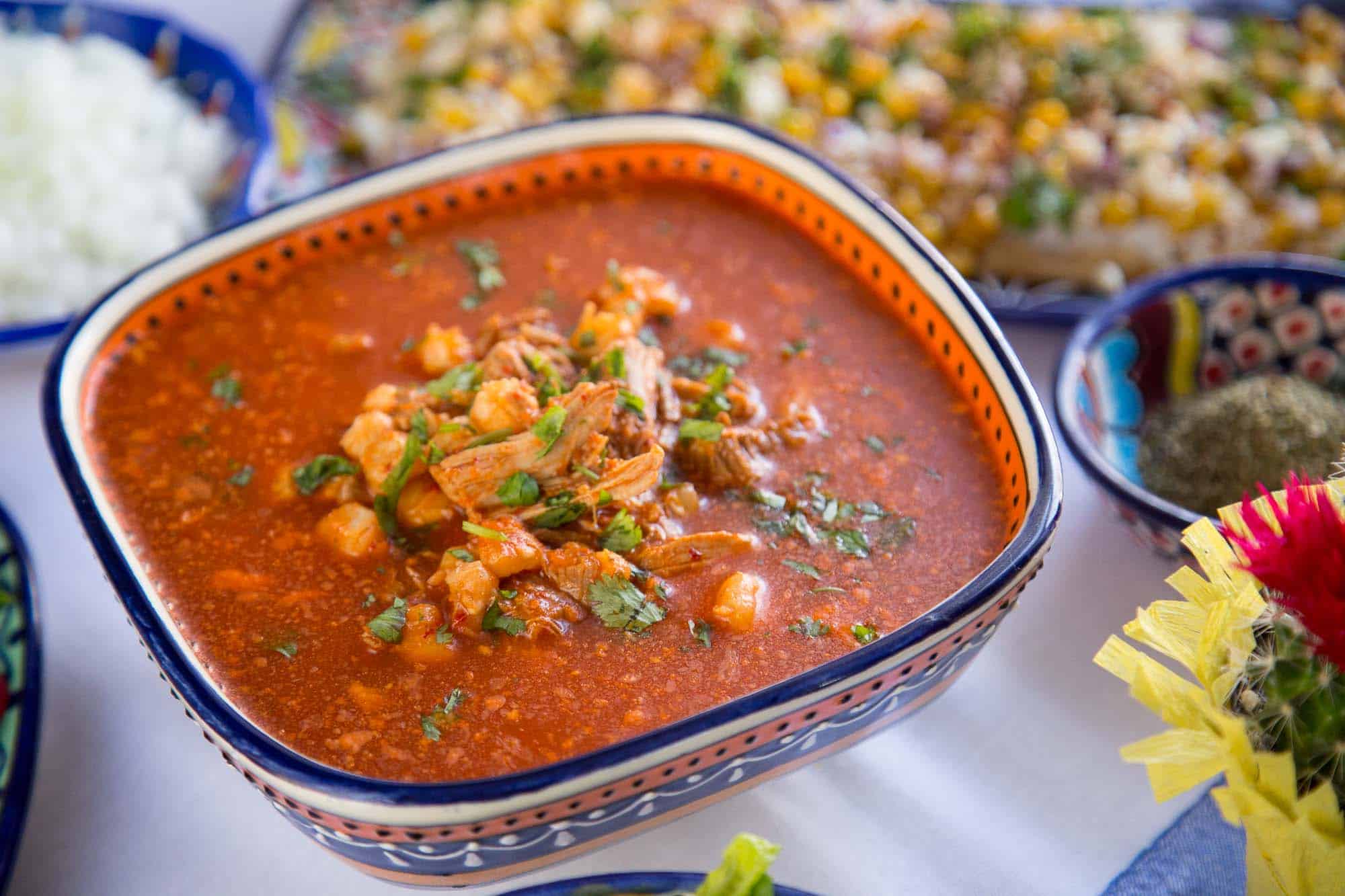 red pozole in a colorful dish on a white tablecloth with other dishes nearby.