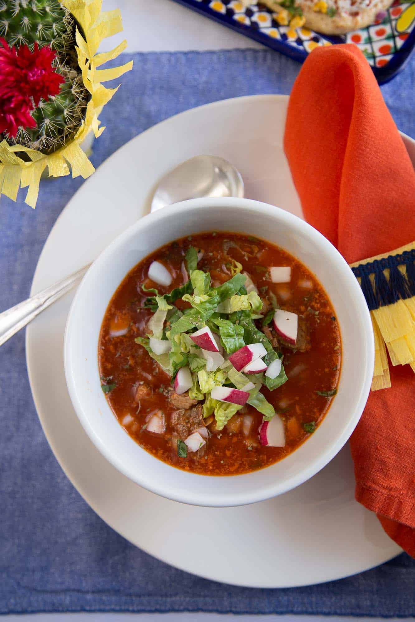 red pozole in a white bowl topped with radishes and romaine.