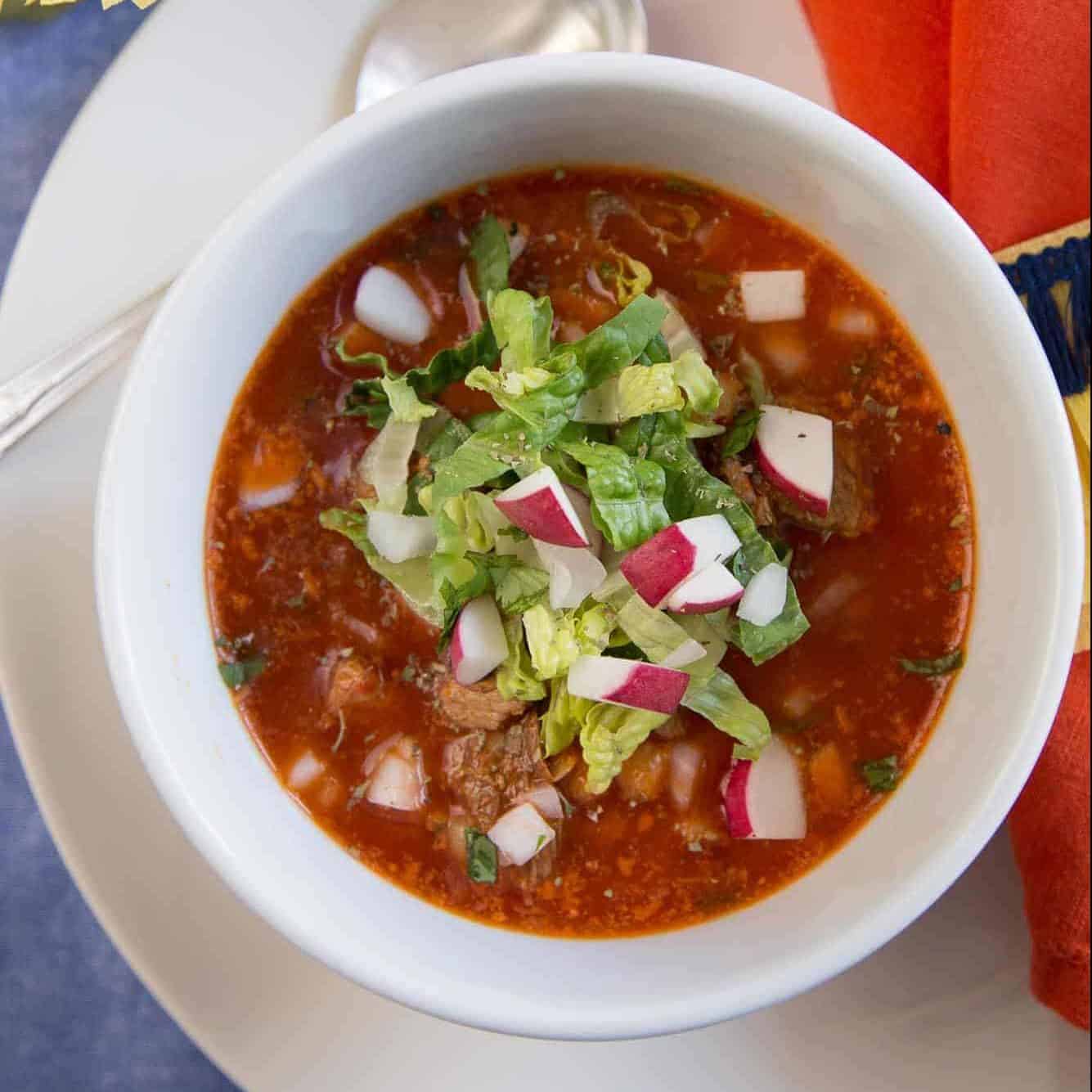red Pozole in white bowl, topped with chopped romaine and chopped radishes.