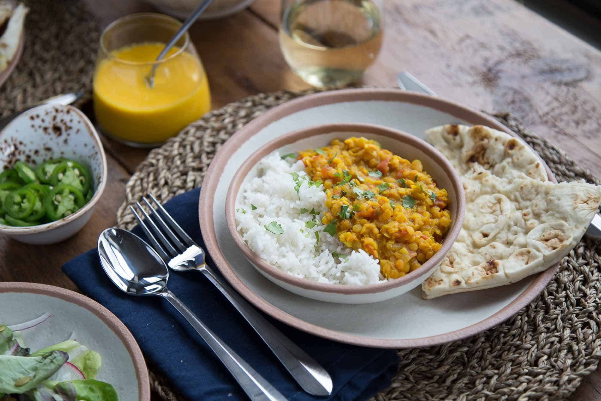 dinner table set with round woven placemats, blue napkins, and bowls of Vegetarian Red Lentil Dal with rice and cilantro.