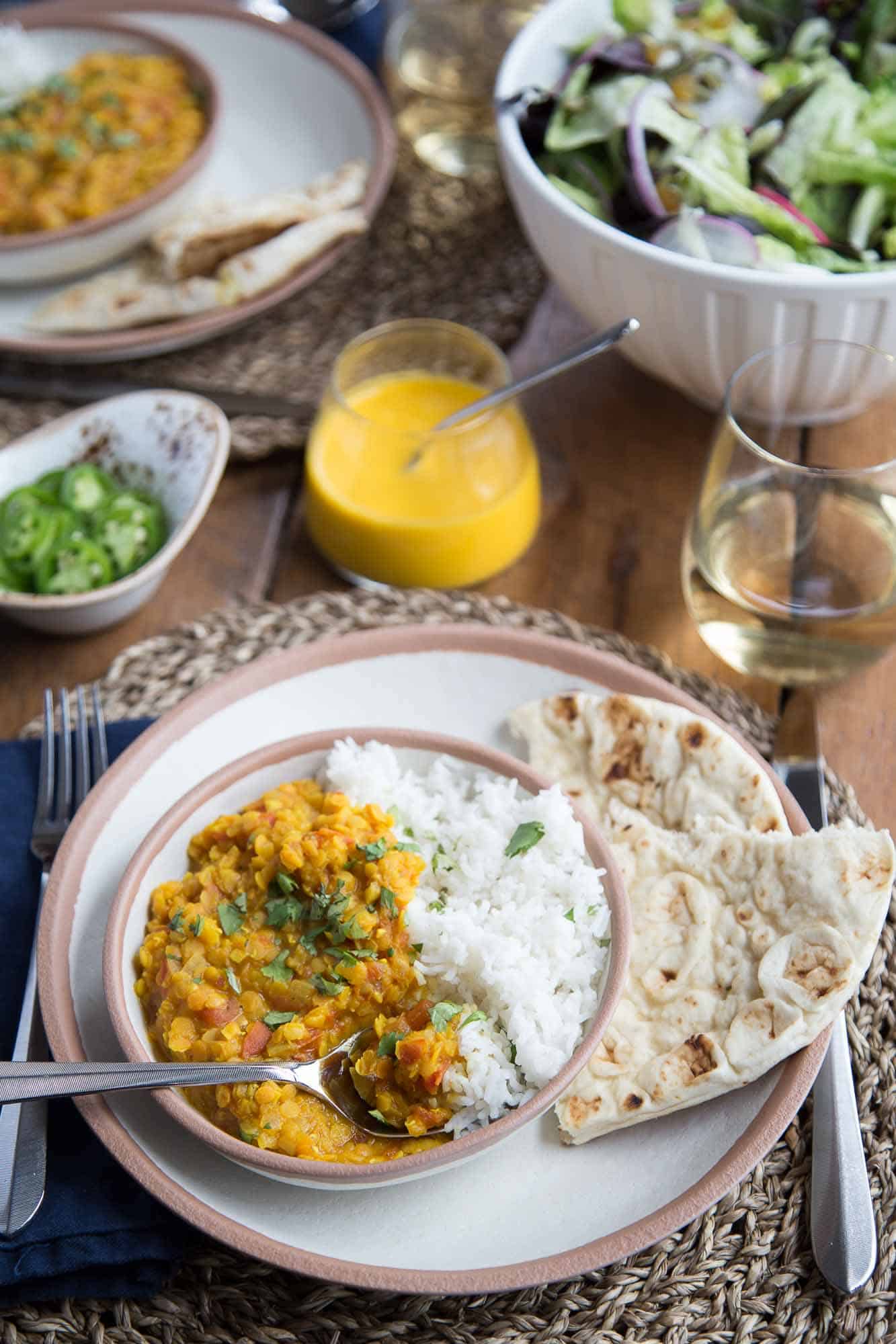 Vegetarian Red Lentil Dal on a dinner table with rice, naan, a green salad, and a glass of white wine.