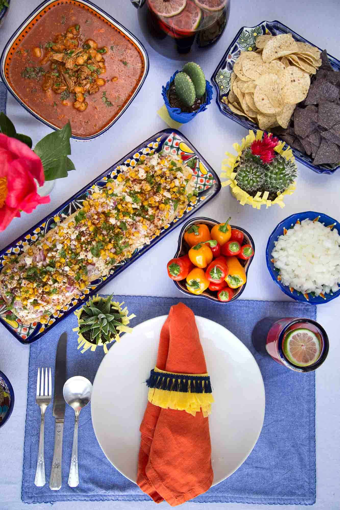 table set for cinco de mayo with colorful mexican dishes, an orange napkin, blue placemat, and cacti decor.