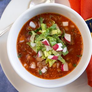 red pozole in a white bowl, topped with chopped romaine and chopped radishes.