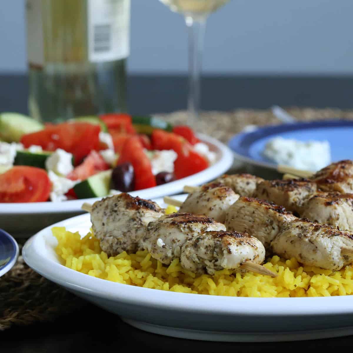 wooden table with chicken kabobs and yellow rice on a platter, and greek salad on another platter.