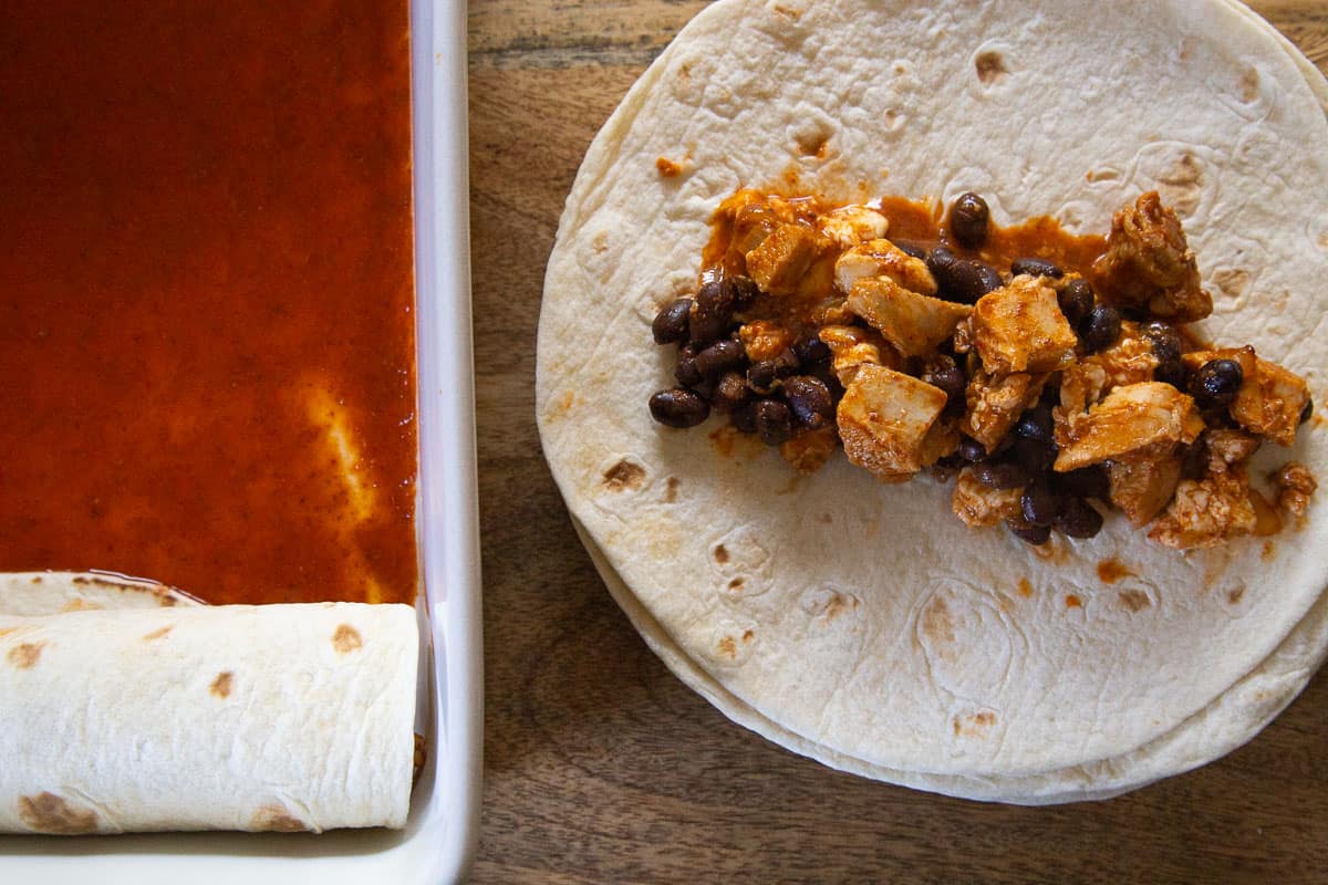 chicken and black beans on a flour tortilla, next to a casserole dish filled with enchilada sauce and a rolled enchilada.