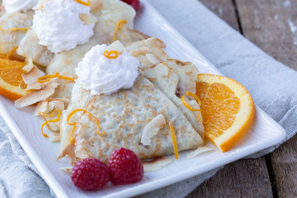 Coconut Milk Crepes on a white platter with raspberries and orange slices.