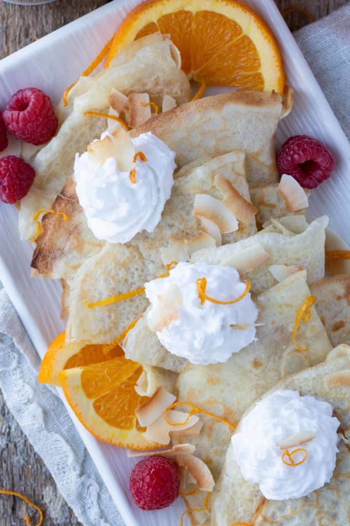 coconut milk crepes stacked on a platter with raspberries, whipped cream, and orange slices.