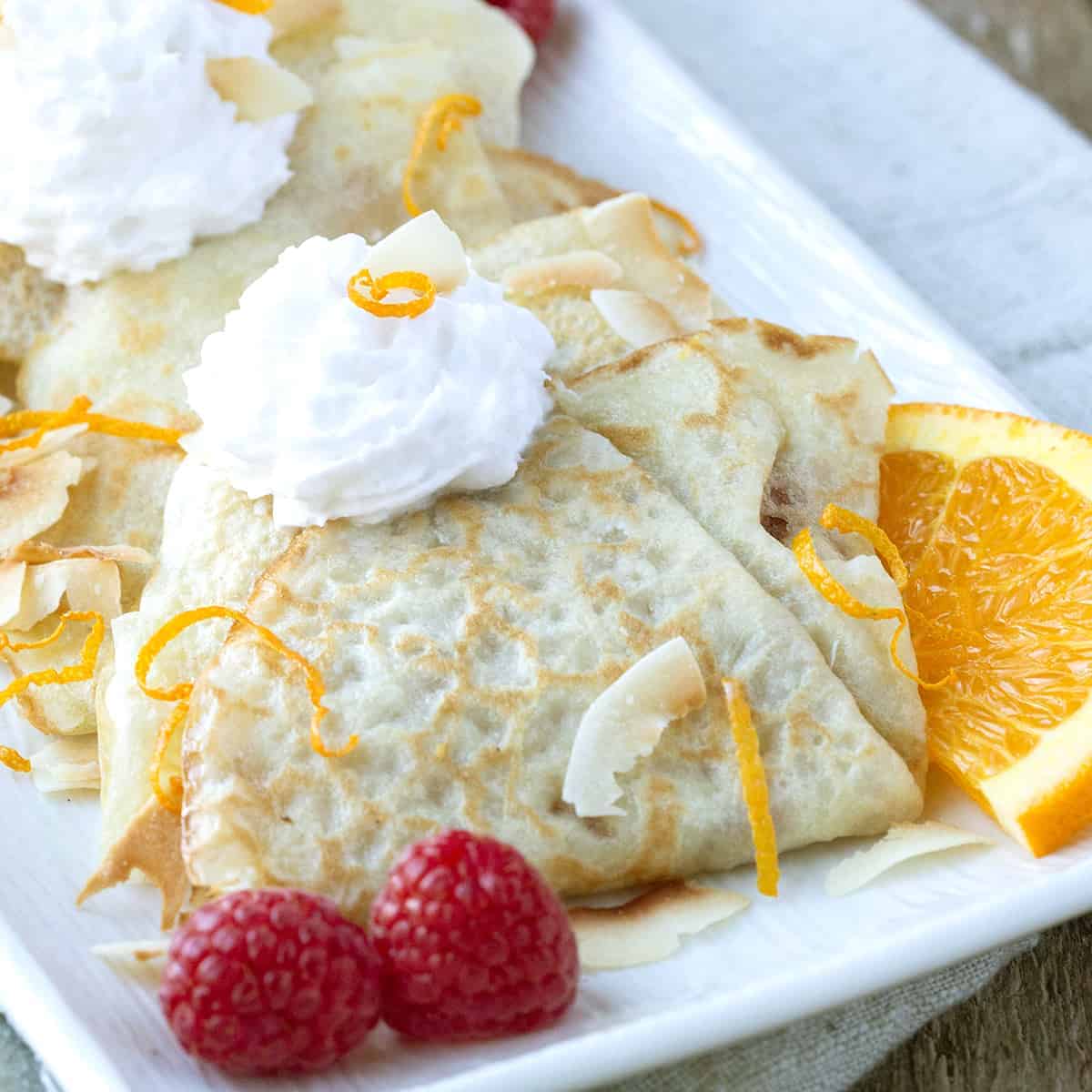 folded crepes on a white rectangular platter, garnished with raspberries, orange slices, and whipped cream.