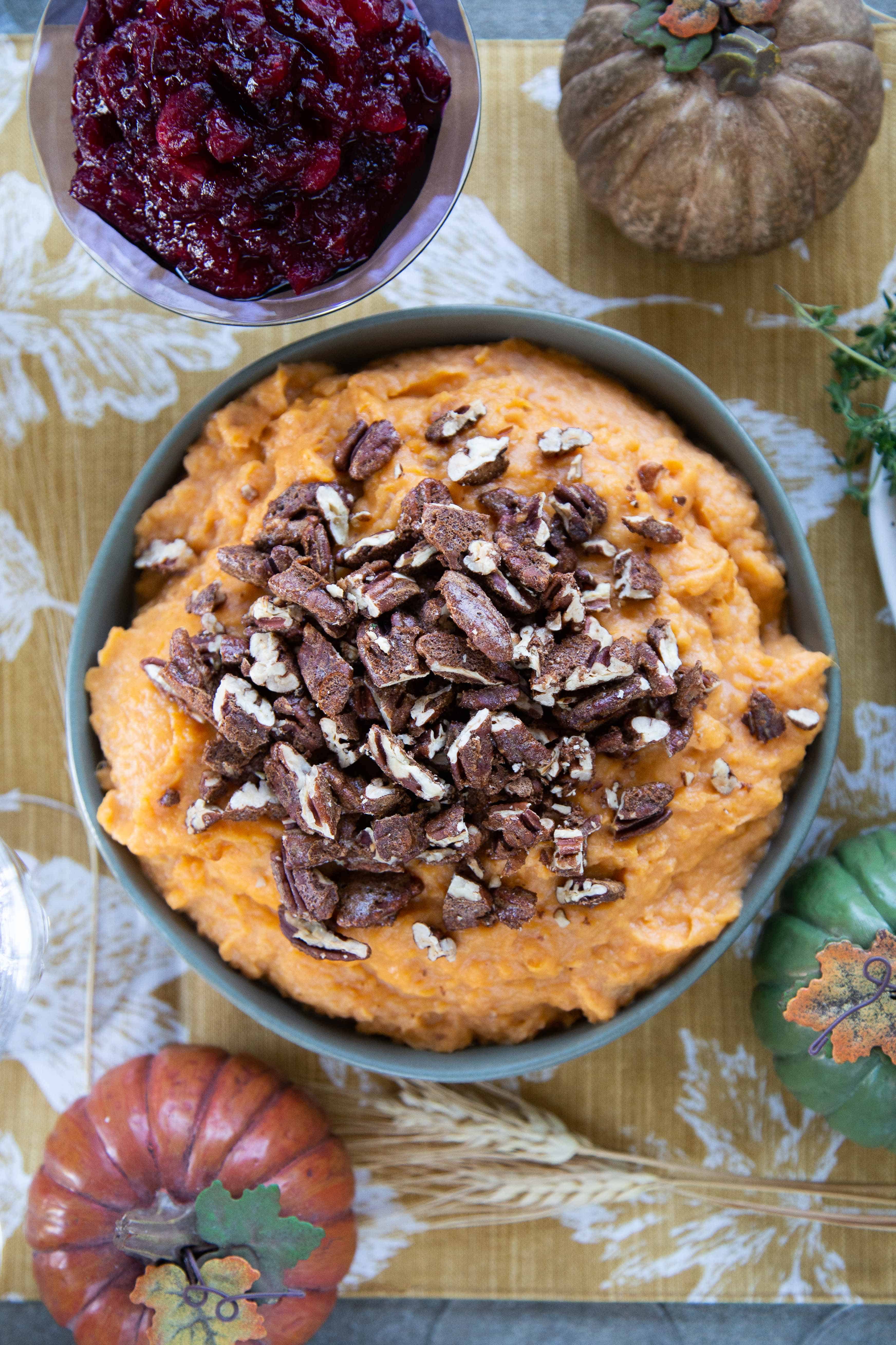 Mashed Sweet Potatoes with Spiced Pecans in a green bowl on a table with cranberry sauce and a gold pumpkin.
