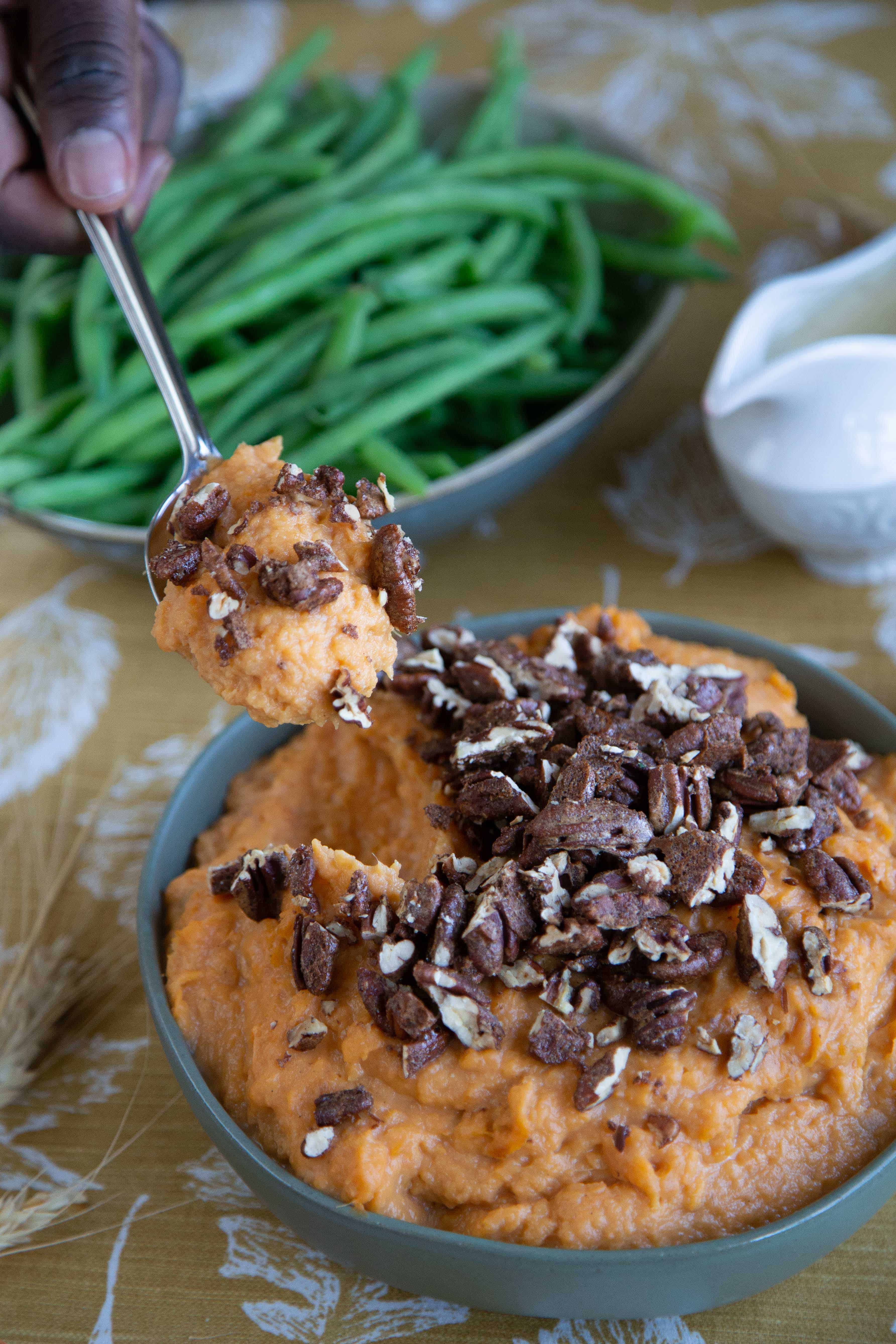 hand holding a spoonful of mashed sweet potatoes topped with spiced pecans on a table with fresh steamed green beans.