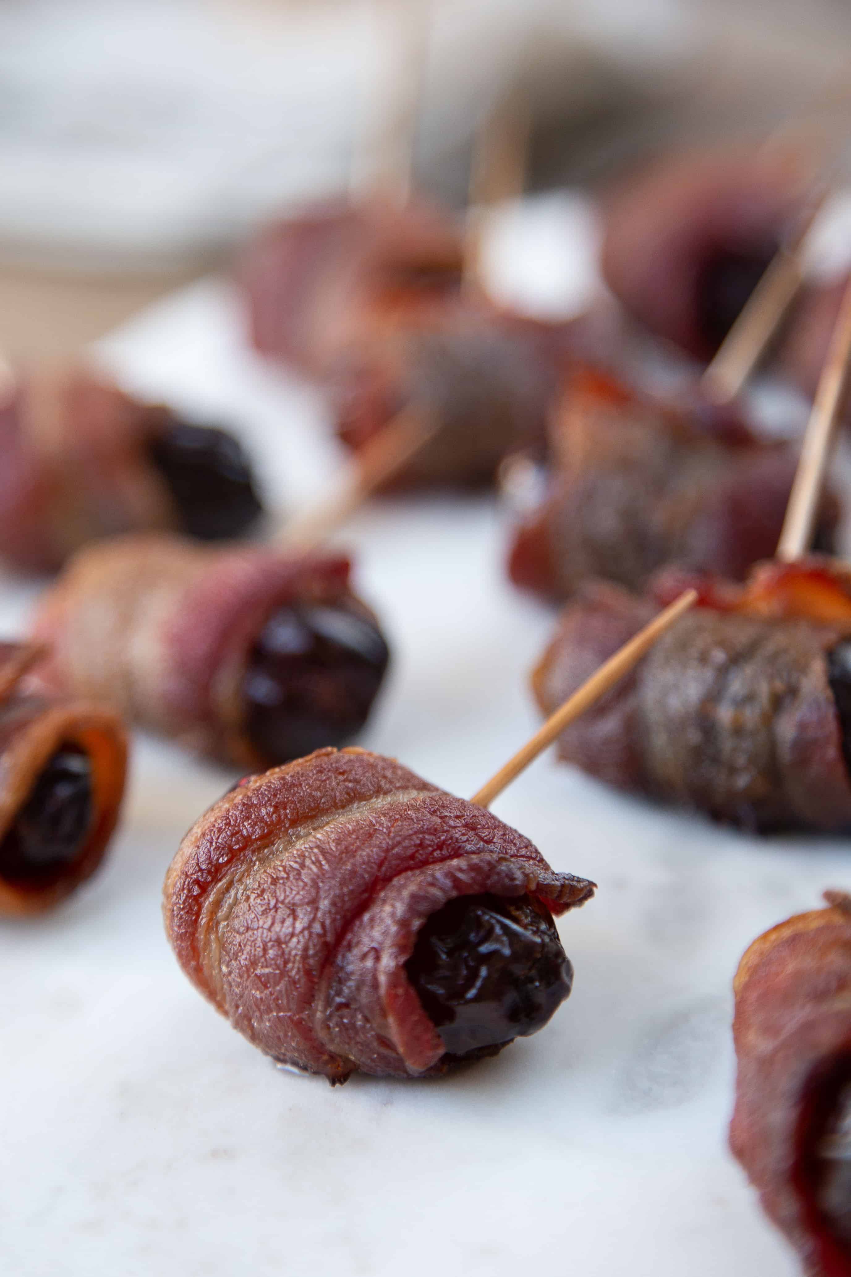 bacon wrapped dates with toothpicks stuck in them, sitting on white parchment paper.