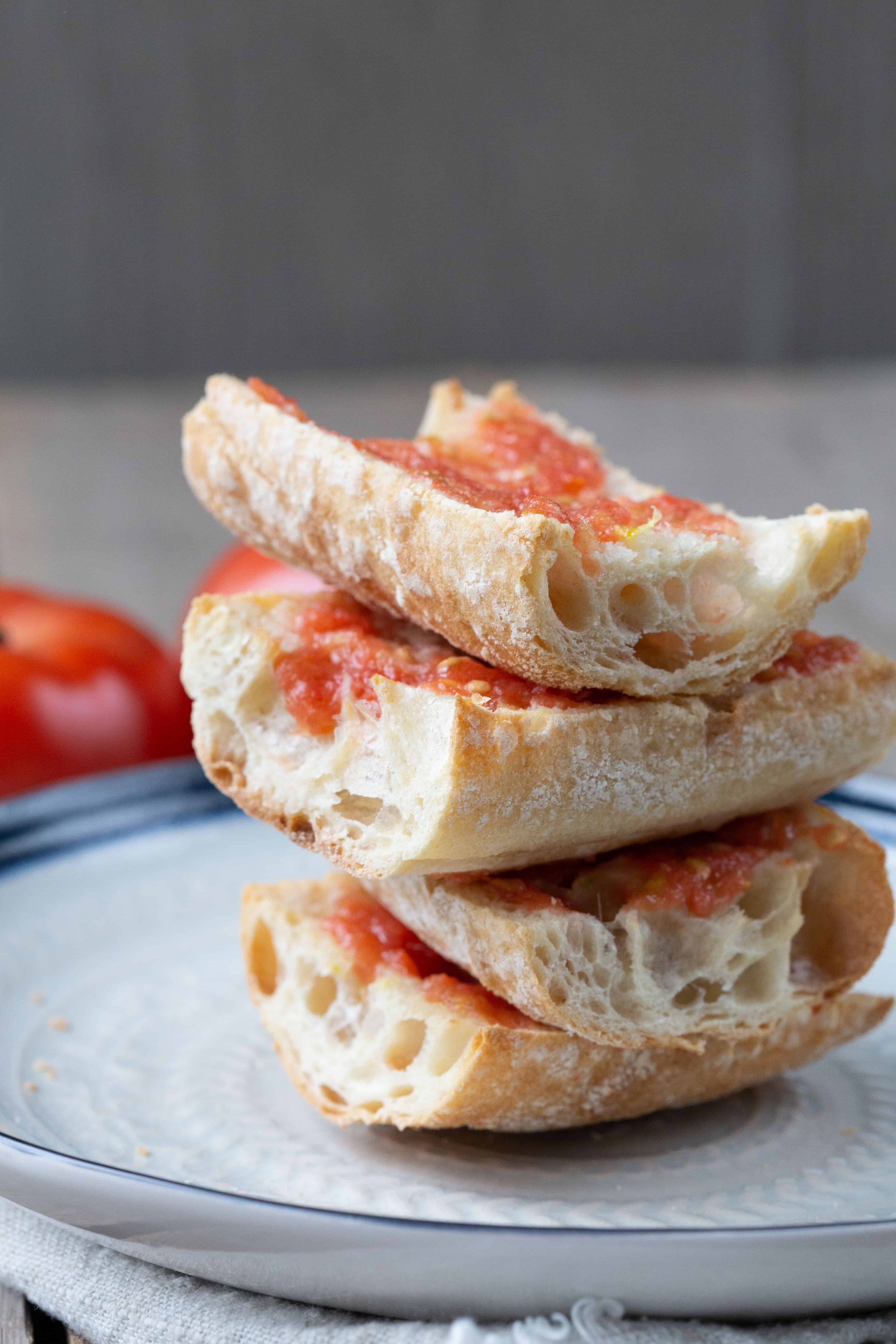 4 pieces of Pan con Tomate stacked on a cream plate with a blue rim.