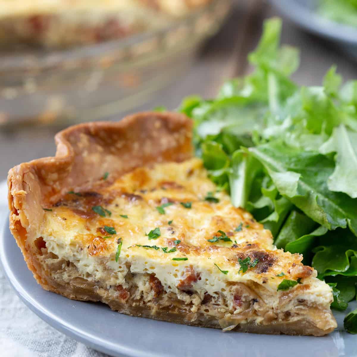 slice of alsatian quiche on a gray plate with a leafy green salad.