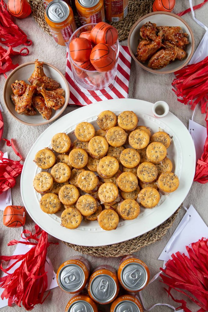 Ritz Crackers Party Sandwiches on a white platter, surrounded by red and white napkins, pompoms, soda, and wings.