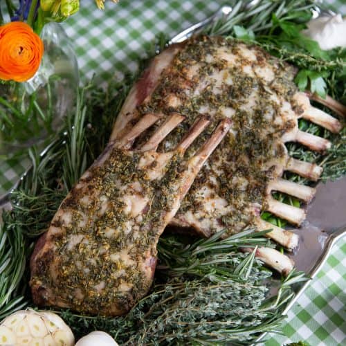 rack of lamb with herbs on a platter, sitting on a green gingham tablecloth.