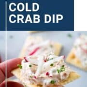 hand holding a saltine cracker loaded with crab dip, radish sticks, and chopped chives.