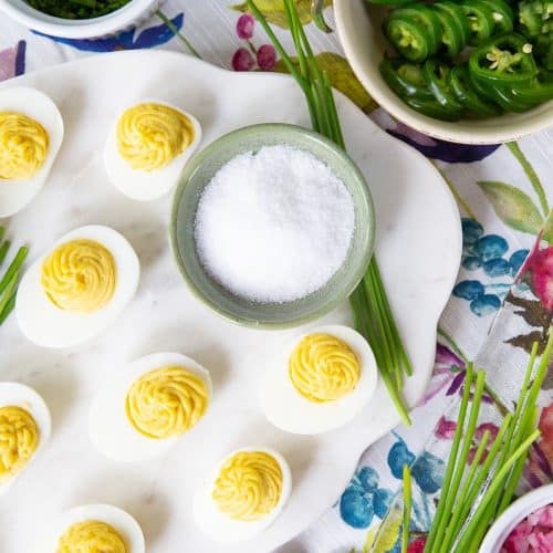 deviled eggs on a white marble tray with a green bowl filled with flaky sea salt.