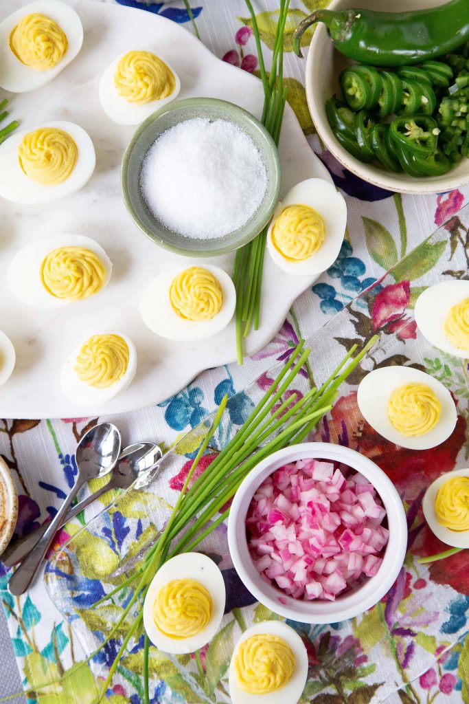 deviled eggs on platters surrounded by chives, sliced jalapeños, and a bowl of flaky sea salt, on a floral tablecloth.