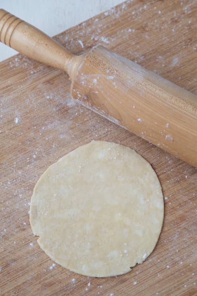 circle of raw empanada dough on a wooden board, next to a rolling pin.