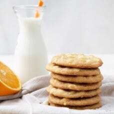 white chocolate cookies stacked up next to a glass of milk and half an orange.