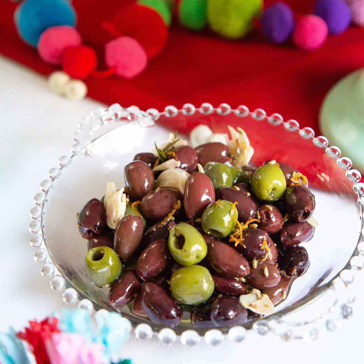 citrus marinated olives in a glass dish on a white tablecloth with a red tablerunner and pom pop decorations.