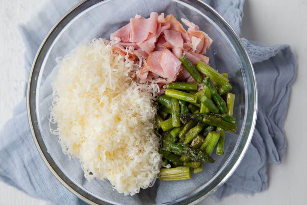 Shredded gruyere cheese, chopped ham, and chopped asparagus in a glass bowl